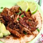 Close up of carnitas on a low carb tortilla with avocado and lime slices and a cilantro garnish.