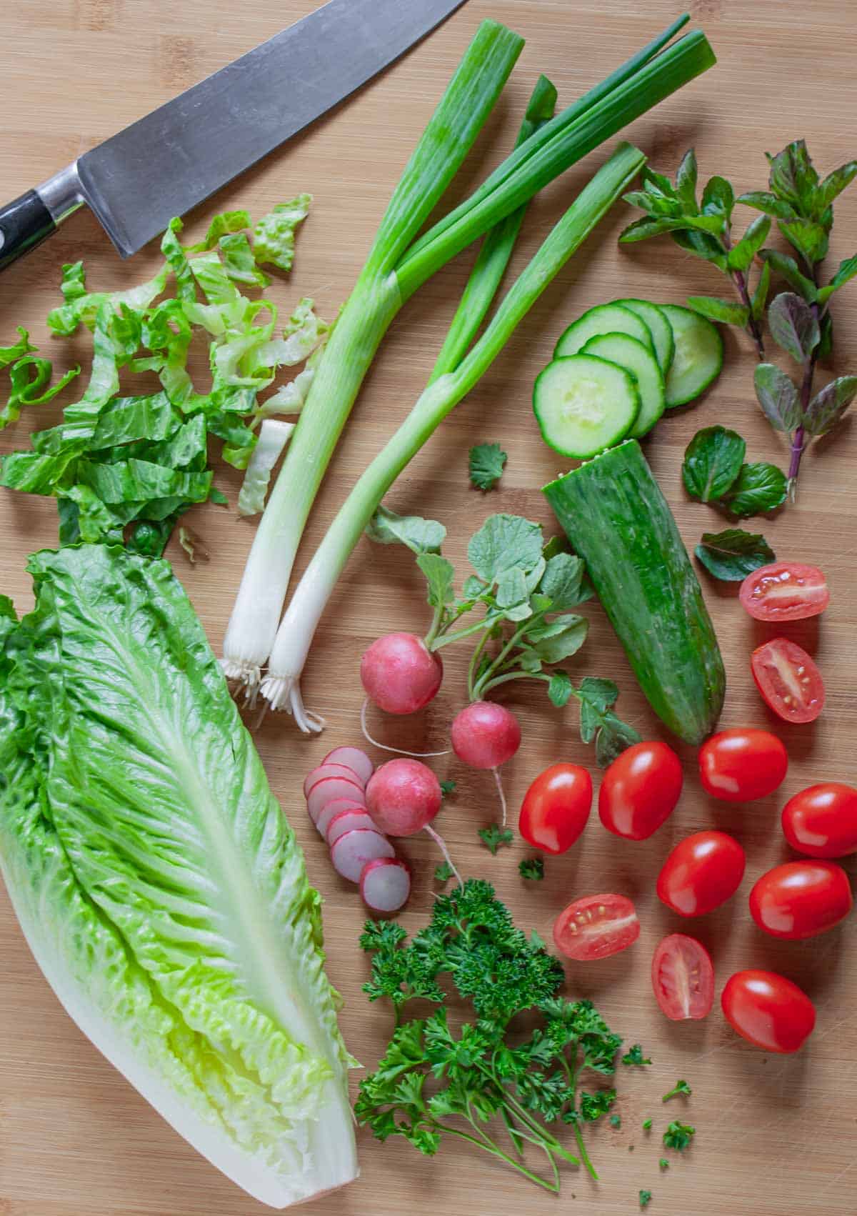 Fresh ingredients for Lebanese fattoush partially chopped on a wood board with a chefs knife.