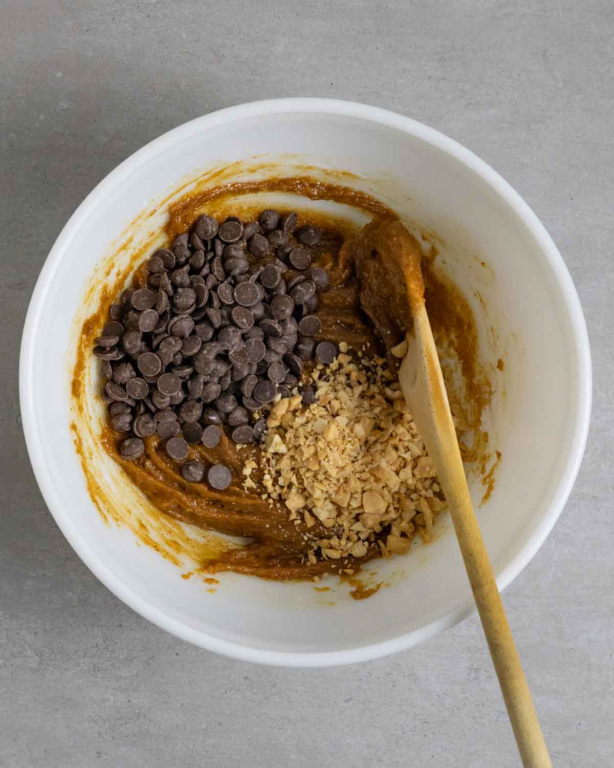 White bowl of cookie batter with a wooden spoon containing piles of chopped peanuts and chocolate chips ready to stir in.