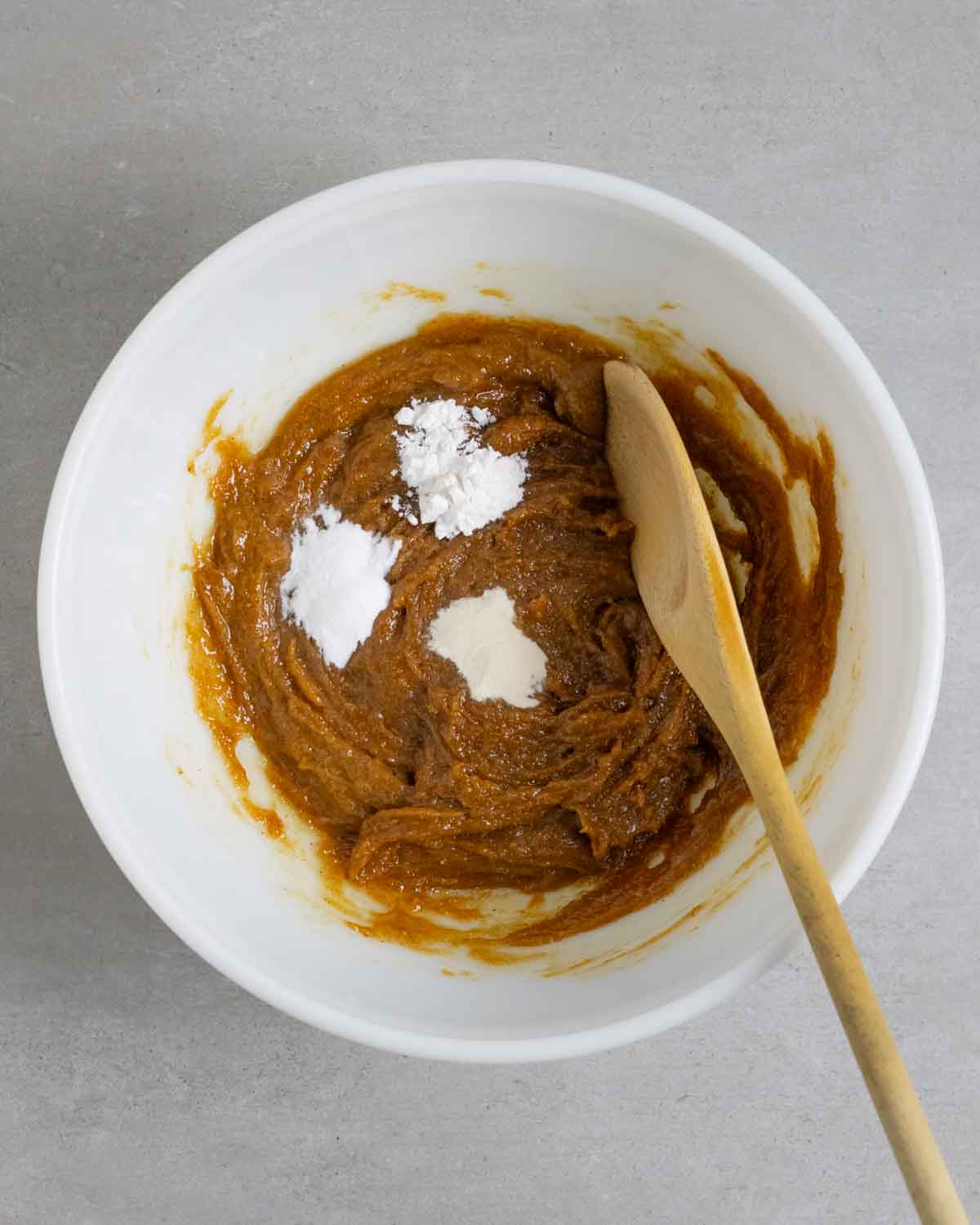 Mixed cookie wet ingredients in a white bowl with a wooden spoon with 3 small piles of baking powder, soda, and xanthan gum ready to mix in.