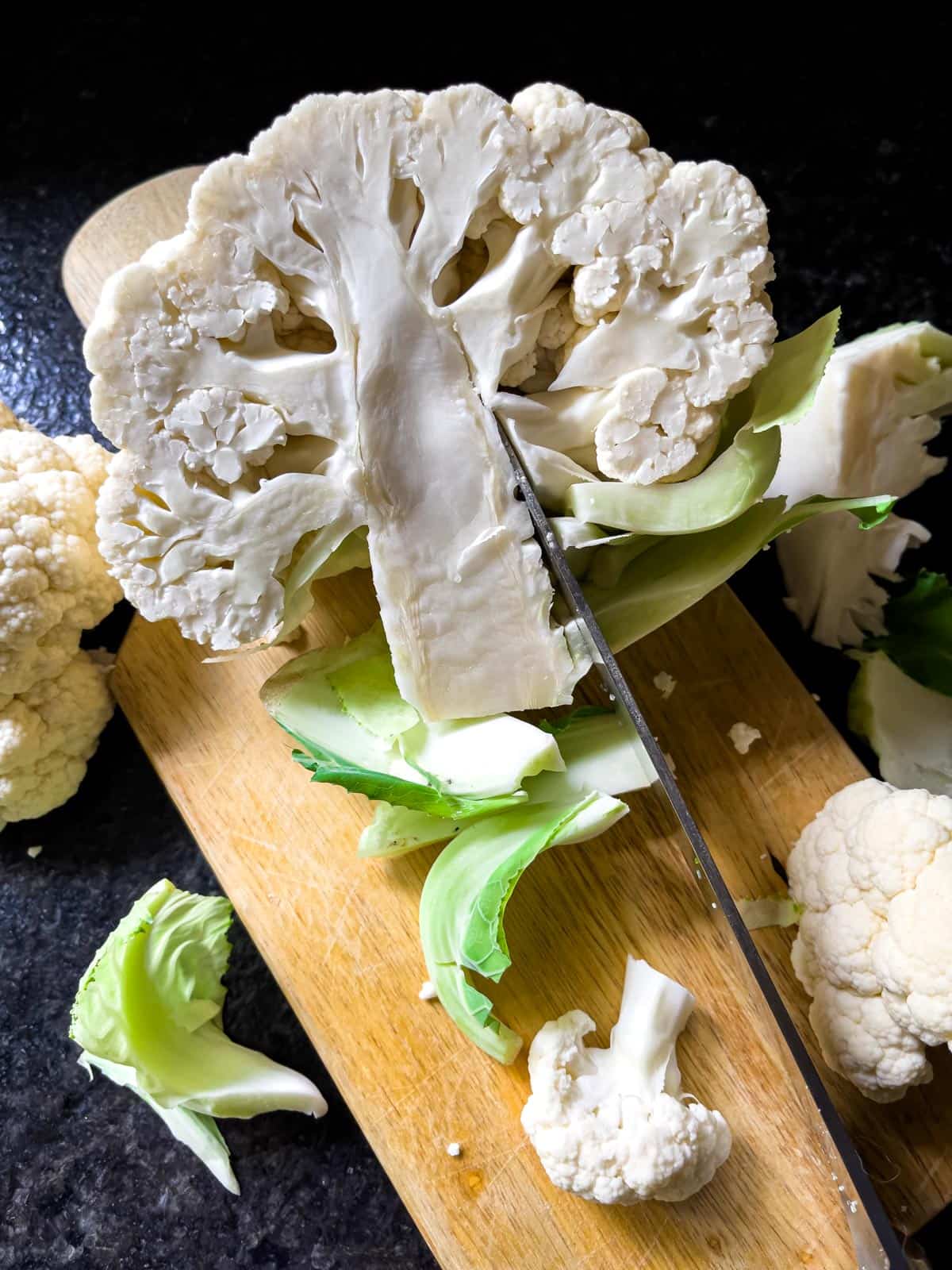 Half a head of fresh cauliflower on a board with a knife cutting the florets away from the core.