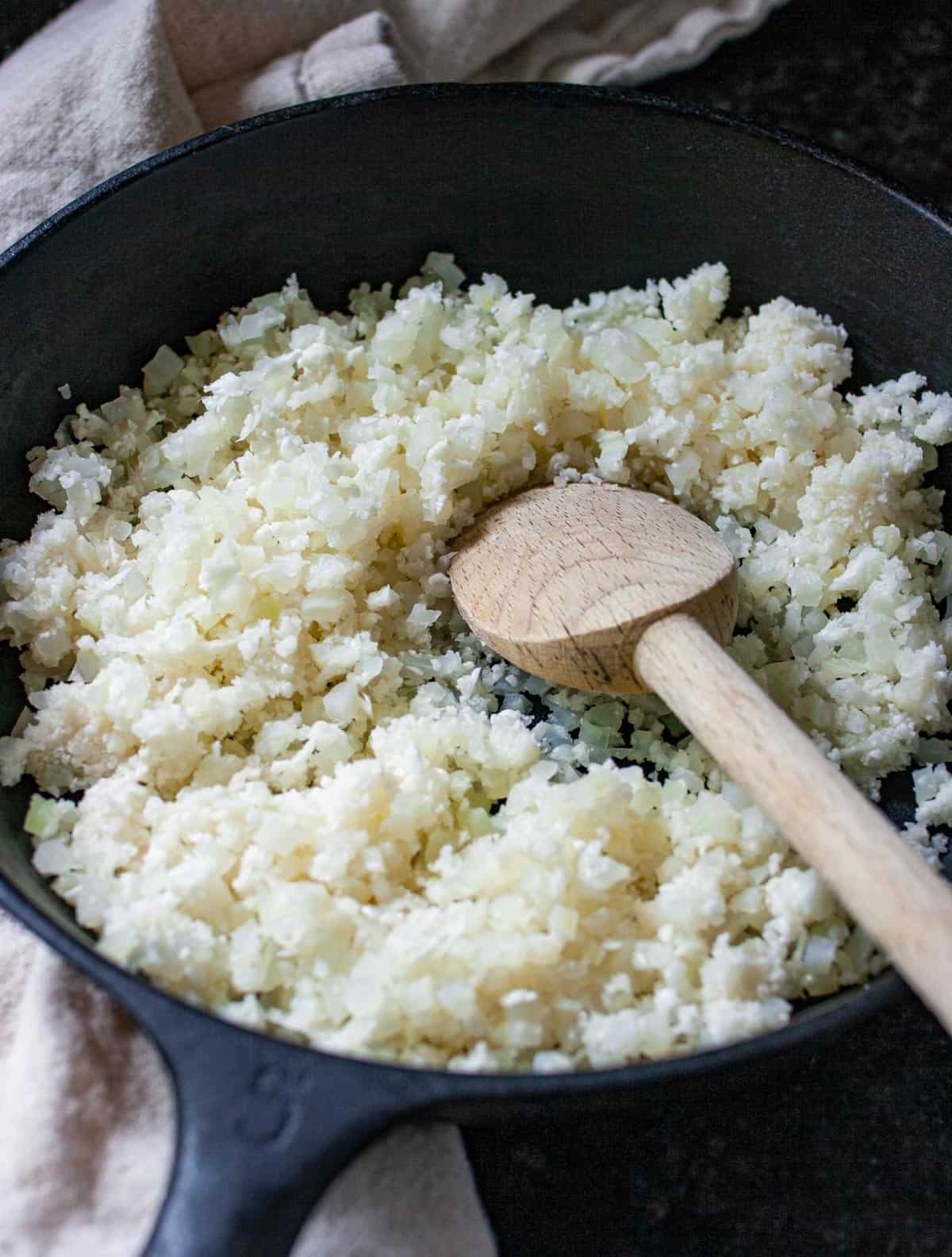 Uncooked fresh riced cauliflower in a cast iron skillet with a wooden spoon in the pan.