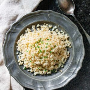 Riced cauliflower on a silver plate with a chopped parsley garnish with a spoon and napkin on the side.