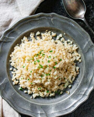Riced cauliflower on a silver plate with a chopped parsley garnish with a spoon and napkin on the side.