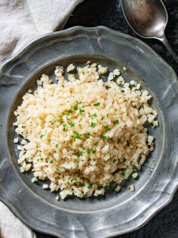 Riced cauliflower on a silver plate with a chopped parsley garnish with a spoon and napkin on the side.