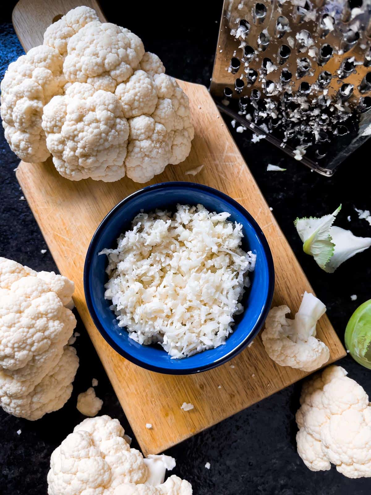 Overhead view of riced fresh cauliflower in a blue bowl on a board with a large floret and a hand grater next to it.