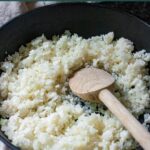 Uncooked fresh riced cauliflower in a cast iron skillet with a wooden spoon in the pan.