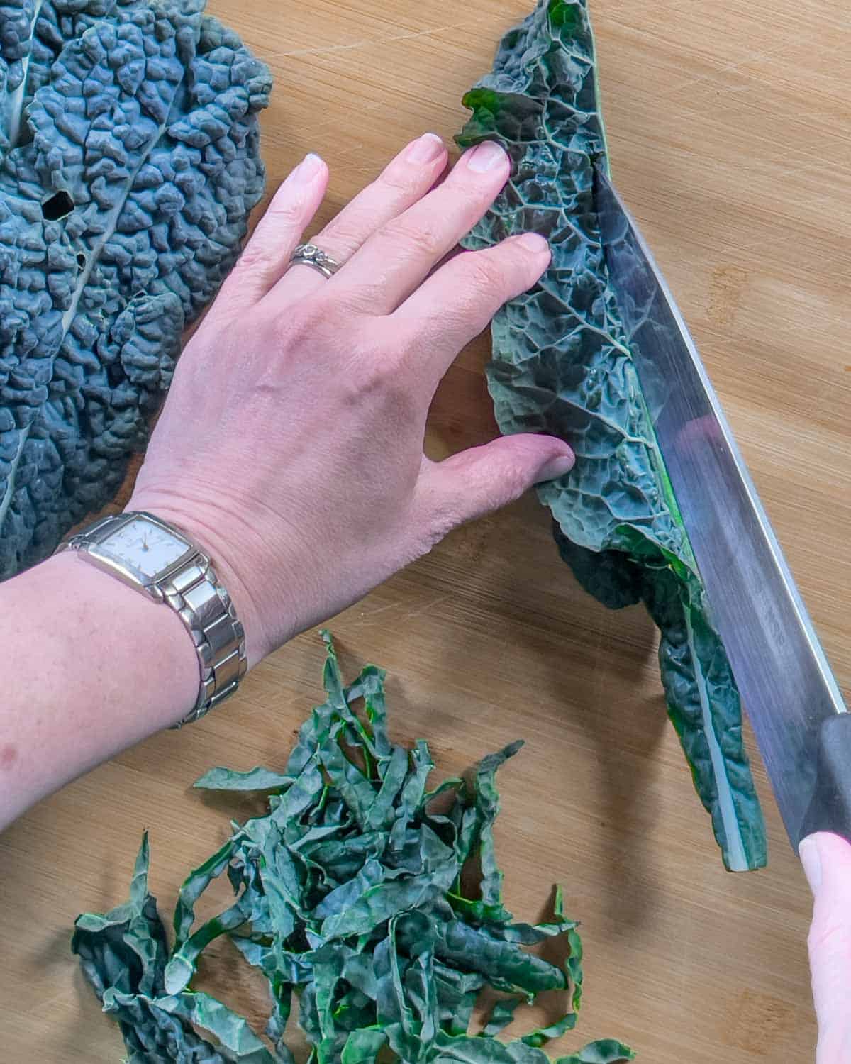 Kale leaves whole and chopped leaves on a cutting board with a hand holding one leaf folded over, slicing the stem out with a knife.