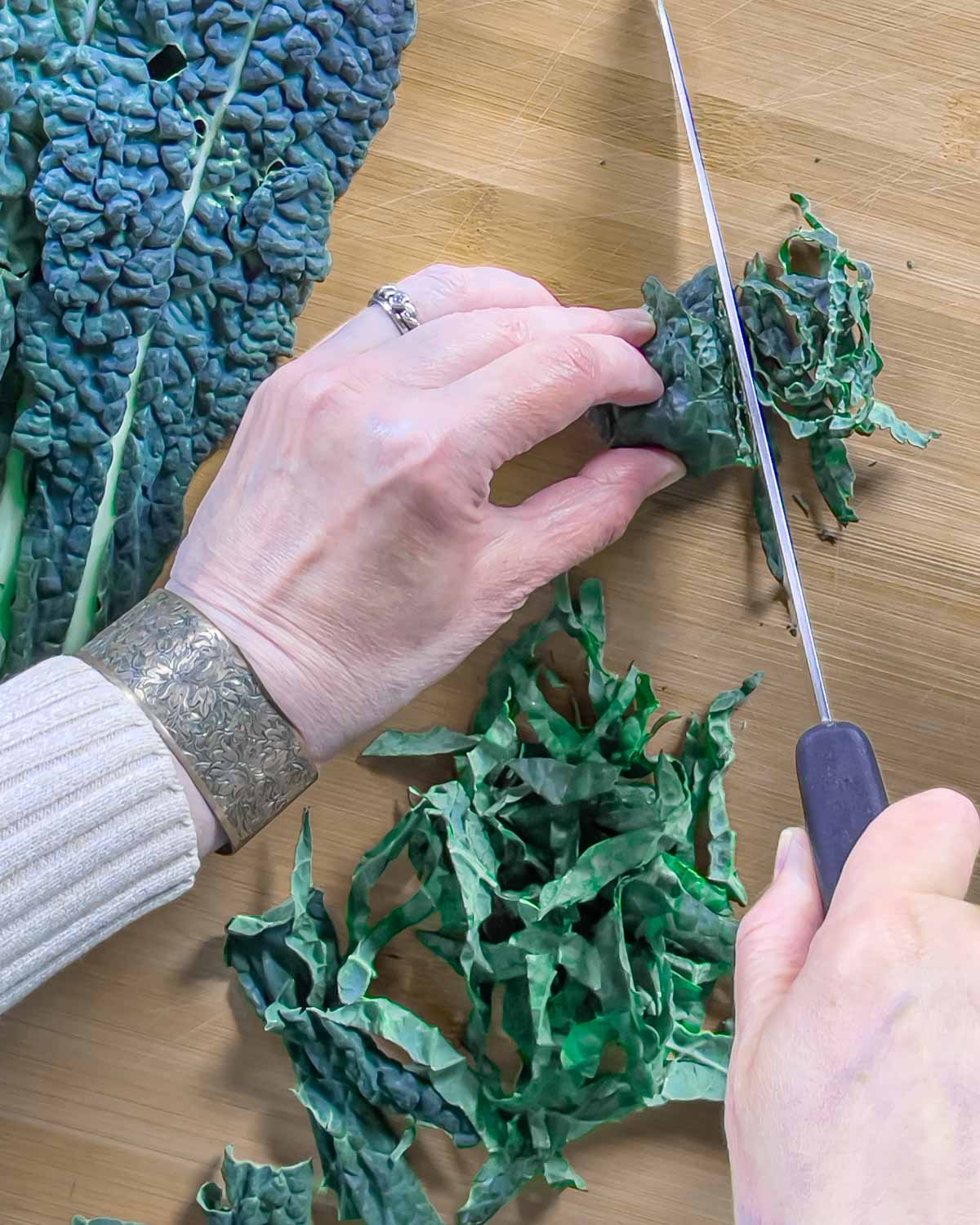 Hands holding kale leaves, in the act of cutting into shreds with a knife.