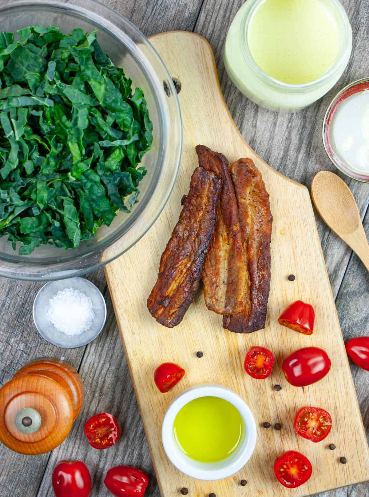 Lacinato kale salad ingredients including a bowl of shredded kale, dressing, cherry tomatoes, bacon, olive oil and salt.