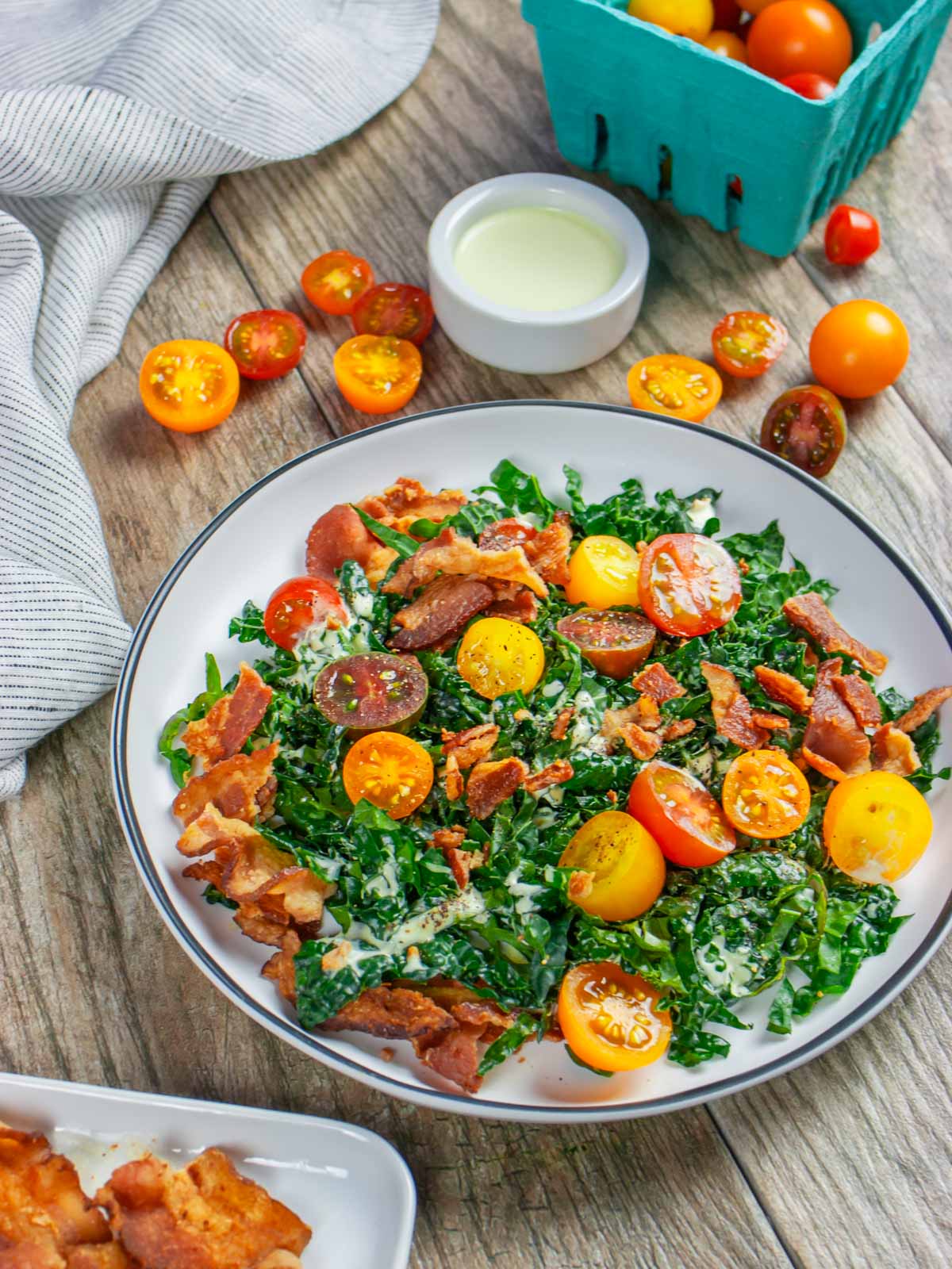 Cavolo nero kale blt salad with orange and red cut cherry tomatoes and bacon. Ramekin of dressing on the side with basket of tomatoes and plate of bacon.