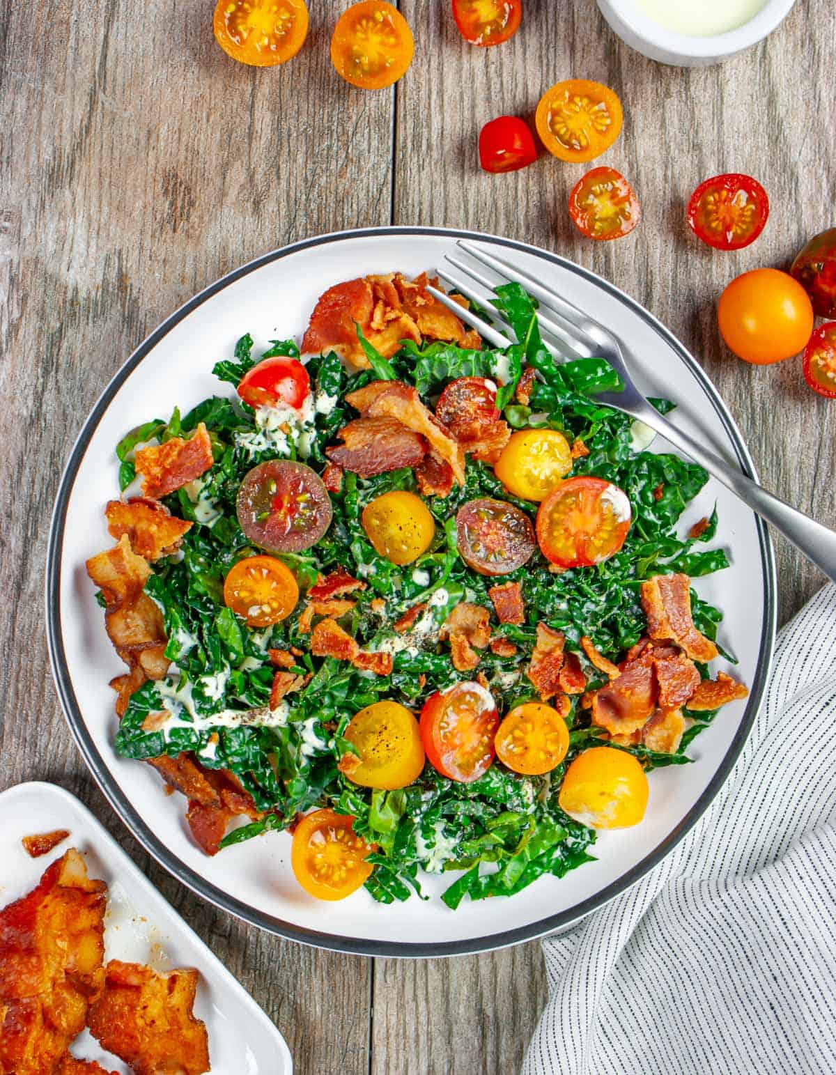 Cavolo nero kale blt salad on a white plate with a fork in it, topped with cut cherry tomatoes and crumbled bacon.