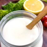 Small mason jar of lemon dressing with a wooden spoon in it, on a board with garnish of parsley, cherry tomato, and a cut in half lemon.