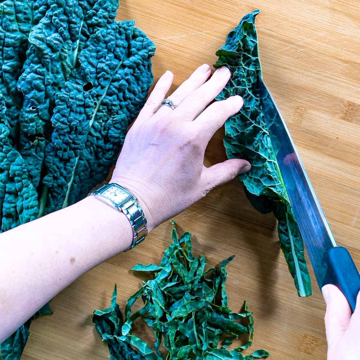 Kale whole and chopped leaves on a cutting board with a hand holding one leaf folded over, slicing the stem out with a knife.