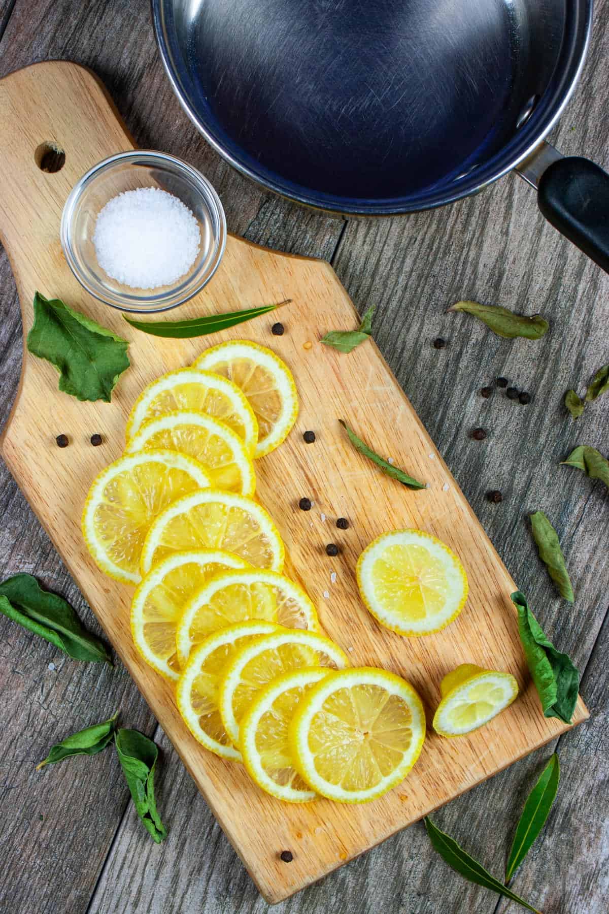 Sliced fresh lemon on a board with peppercorns and spices, some salt in a ramekin, all on a grey wood counter with a sauce pan next to it.