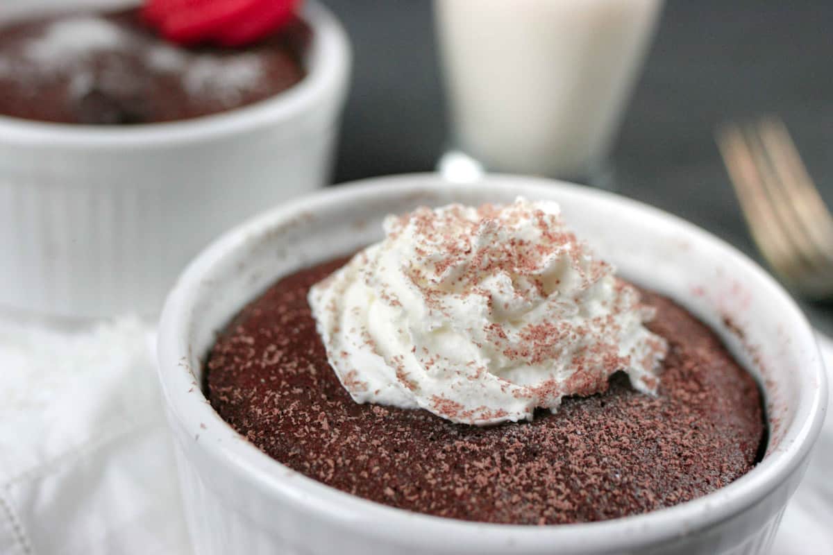Two chocolate mug cakes in white ramekins with whipped cream and shaved chocolate on top.