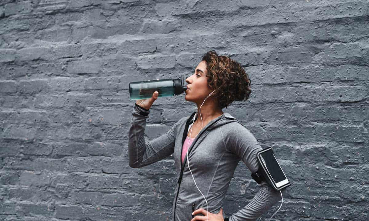 Woman drinking from a sport water bottle in grey sweats with a cell phone strapped to her upper arm with earbuds.