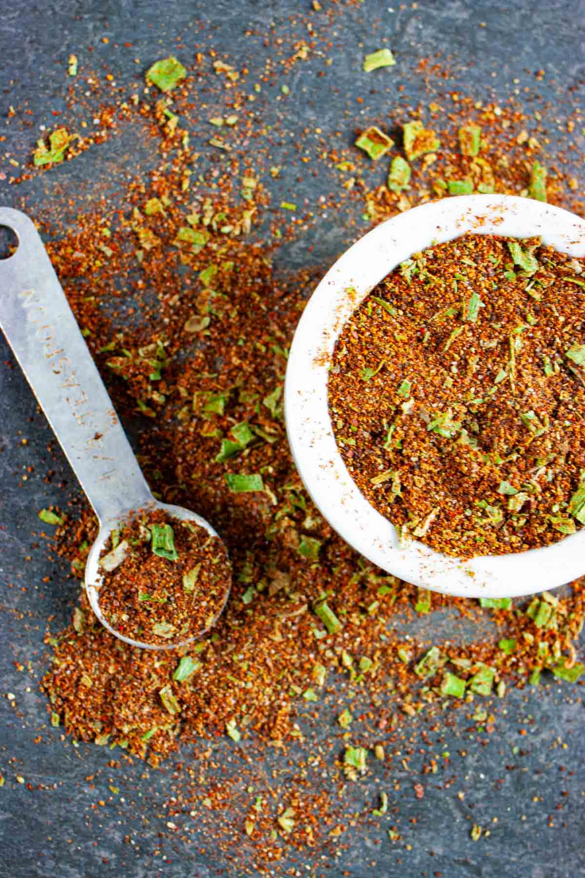 Taco seasoning in a small white bowl and teaspoon and scattered on a grey background.