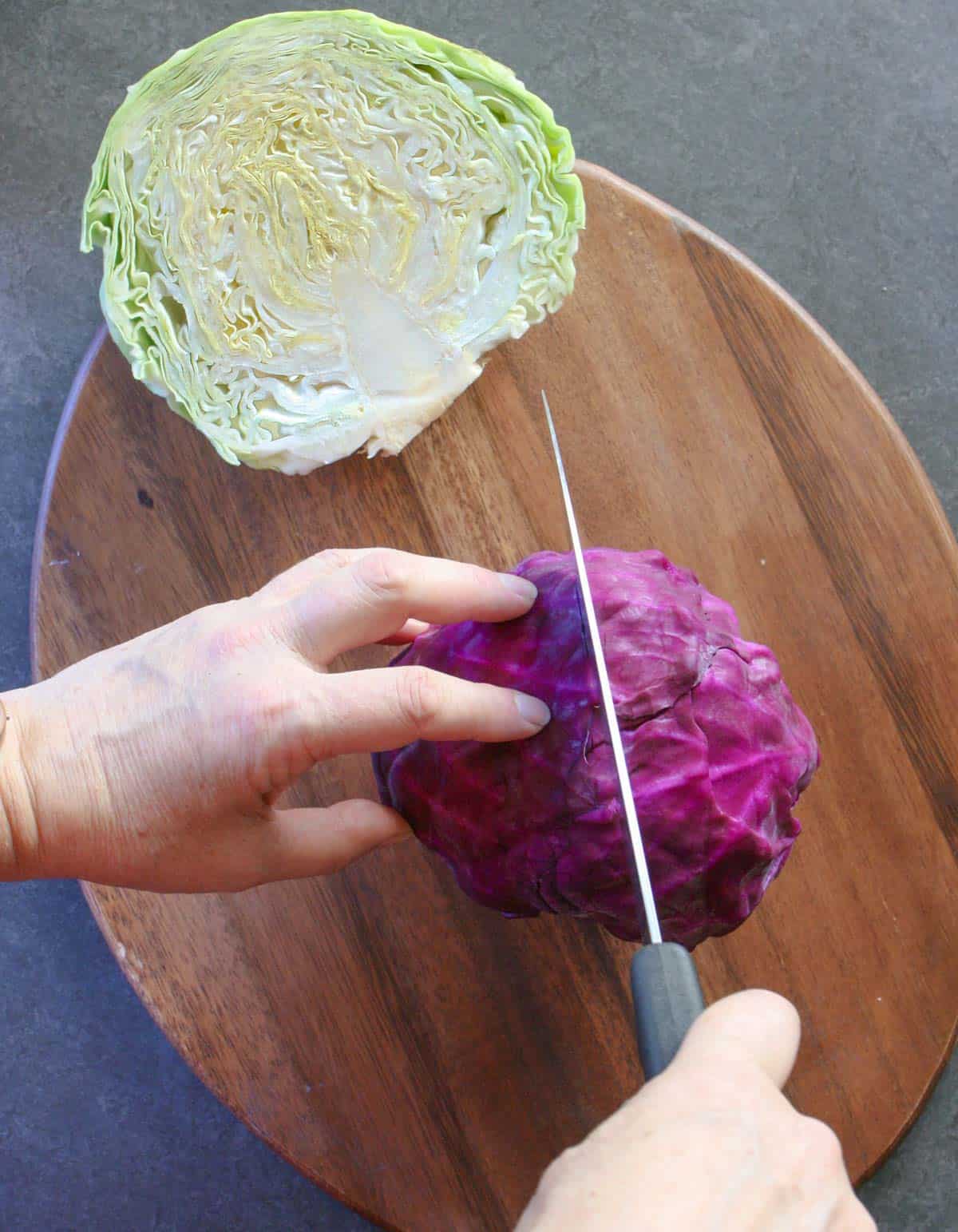 Half a green cabbage on a cutting board with a person cutting a whole red cabbage in half with a knife.