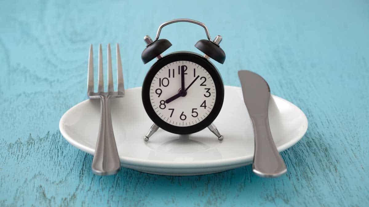 White plate with knife and fork with an alarm clock between them on a blue background.