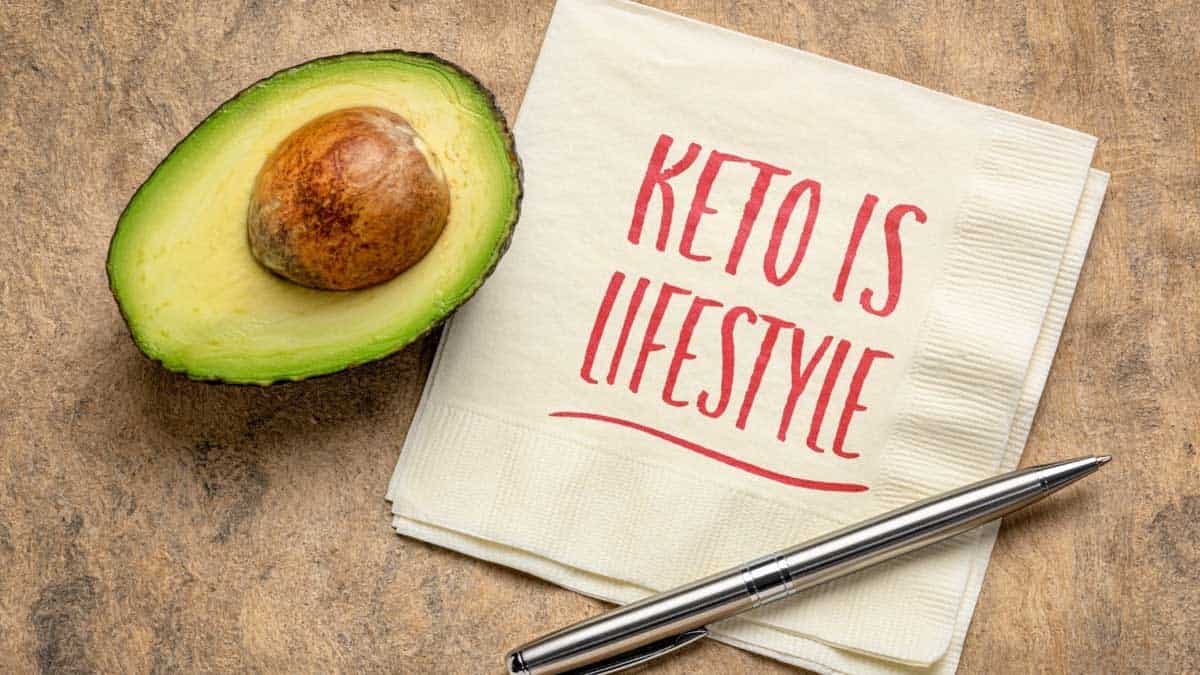 Avocado half with pit on a brown background next to a square paper napkin with a pen on it and the words Keto is Lifestyle written in red on the napkin.