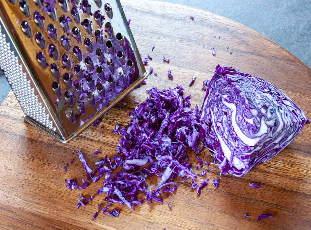 Red cabbage quarter on a wood cutting board with some grated next to a box or hand grater.