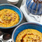 Two bowls of porridge topped with syrup, pumpkin seeds, and pecans with decorative white cup of syrup on the side and a blue blown glass pumpkin.