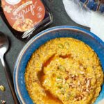 Bowl of porridge with a spoon next to it, bottle of maple pecan syrup laying down, and part of a blue glass pumpkin.