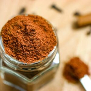 Glass spice jar with sugar-free pumpkin spice seasoning, a measuring spoon full of spice, and whole cloves and cinnamon sticks as garnish.