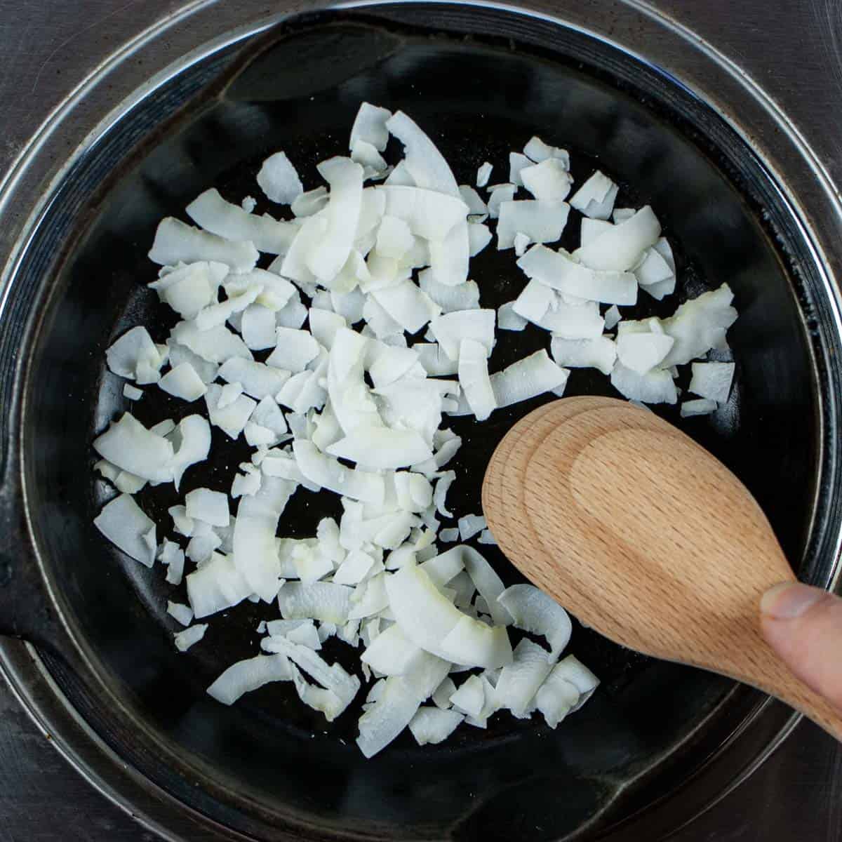 Raw coconut flakes being stirred in a small cast iron frying pan.