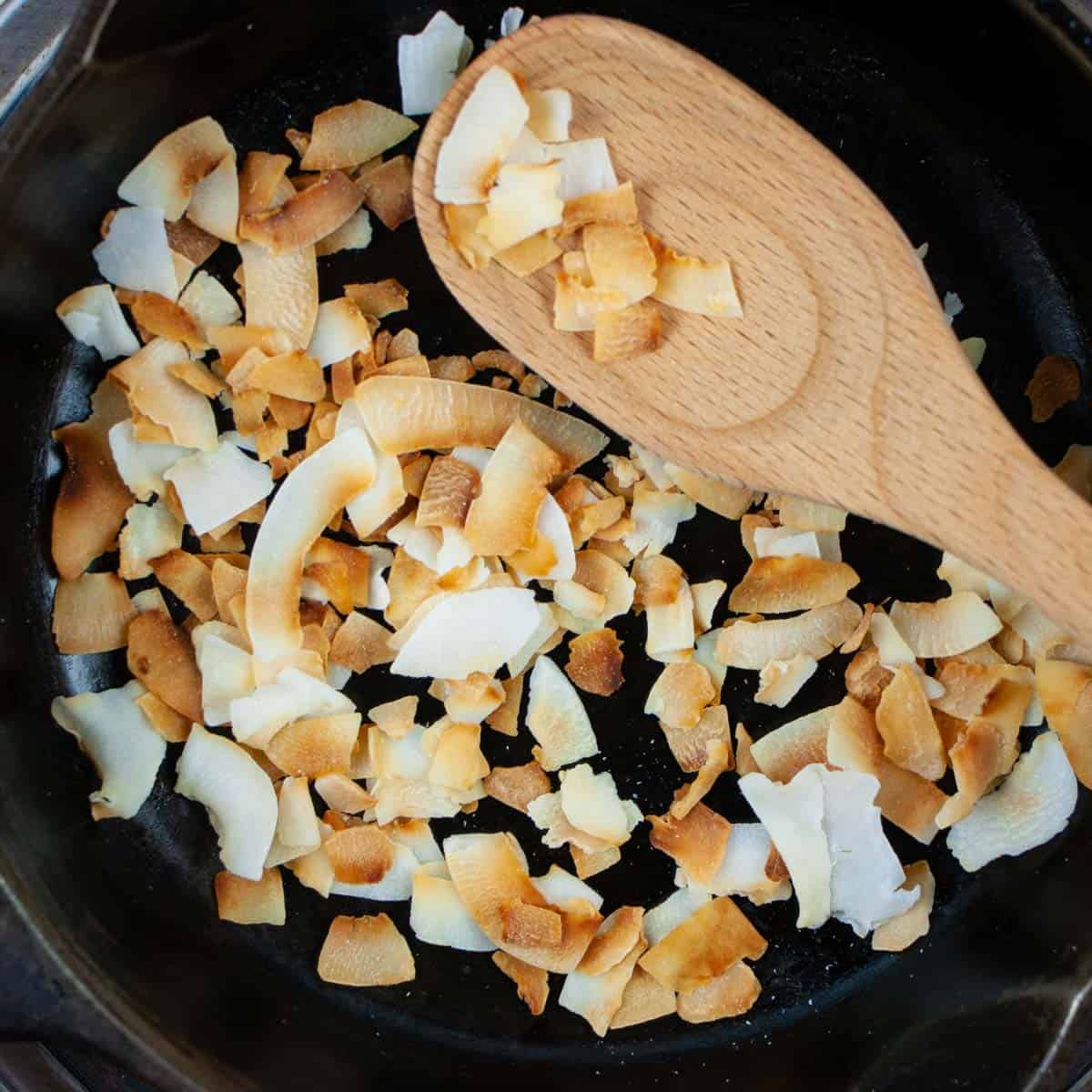 Toasted coconut flakes in a cast iron pan with some on a wooden spoon.