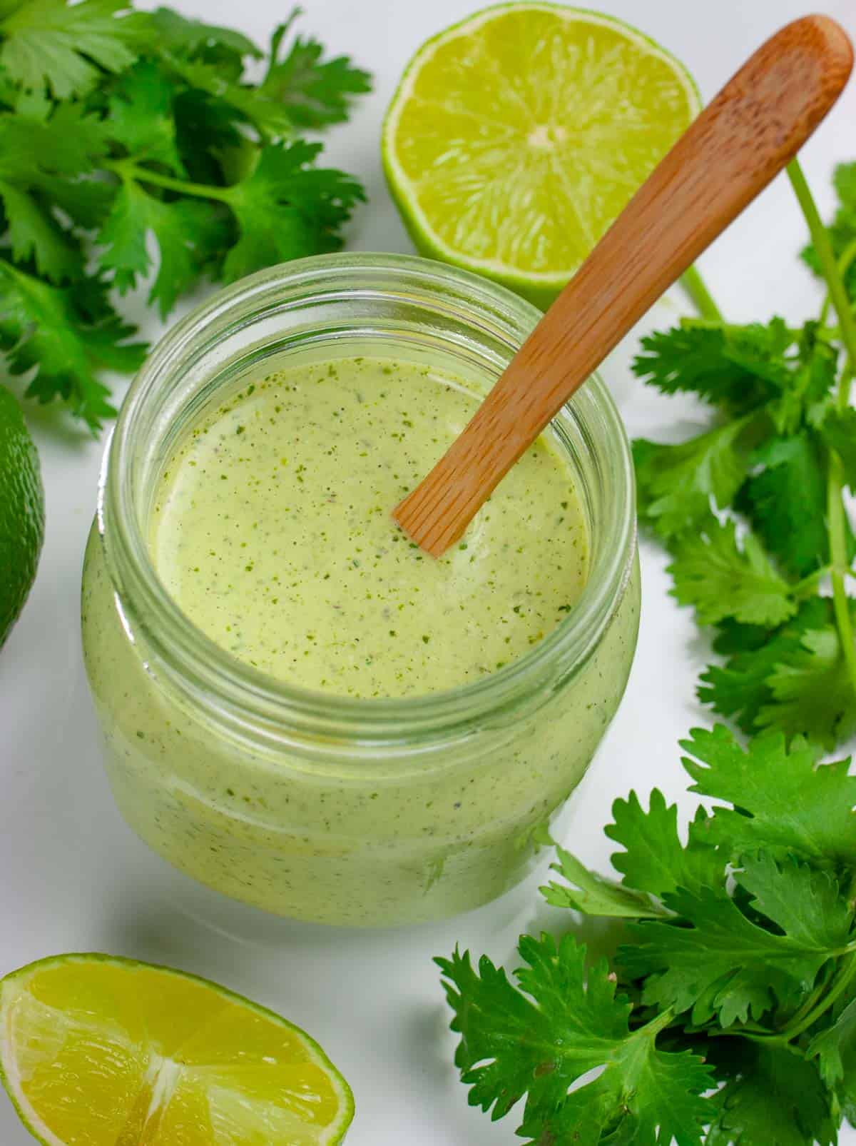 Wide mouth small jar of light green cilantro lime dressing with a small wooden spoon in it, with cilantro and lime halves surroounding the jar.
