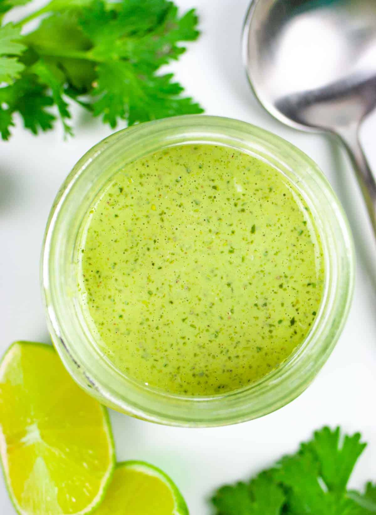 Looking down on a wide mouth jar of dressing on a white cloth with a spoon, lime wedges, and sprigs of cilantro.