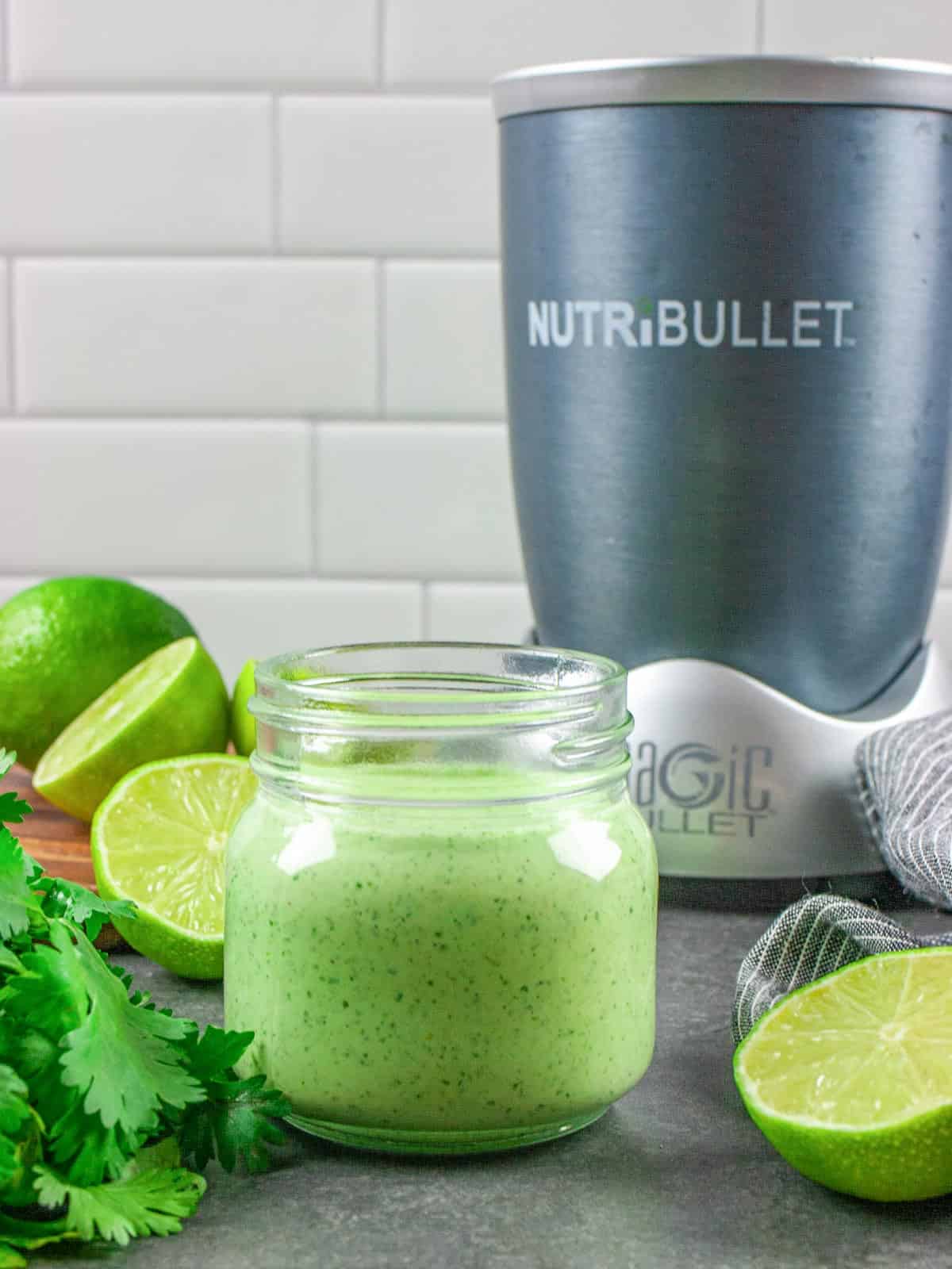 Wide mouth jar of creamy dressing sitting in front of a nutribullet blender with some lime halves and cilantro surrounding the jar.