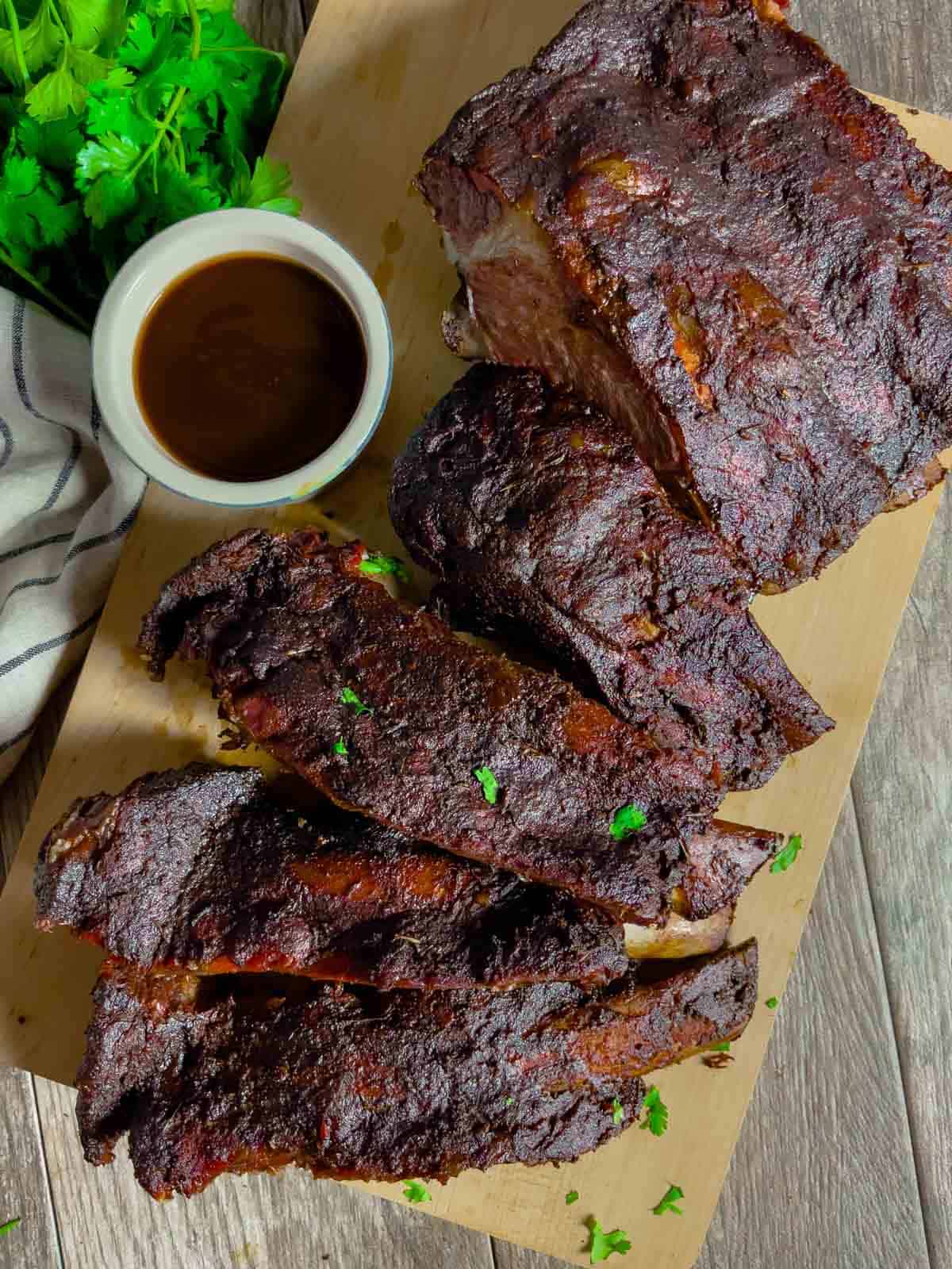 Rack of smoked beef ribs sliced and stacked on a board with ramekin of barbeque sauce.