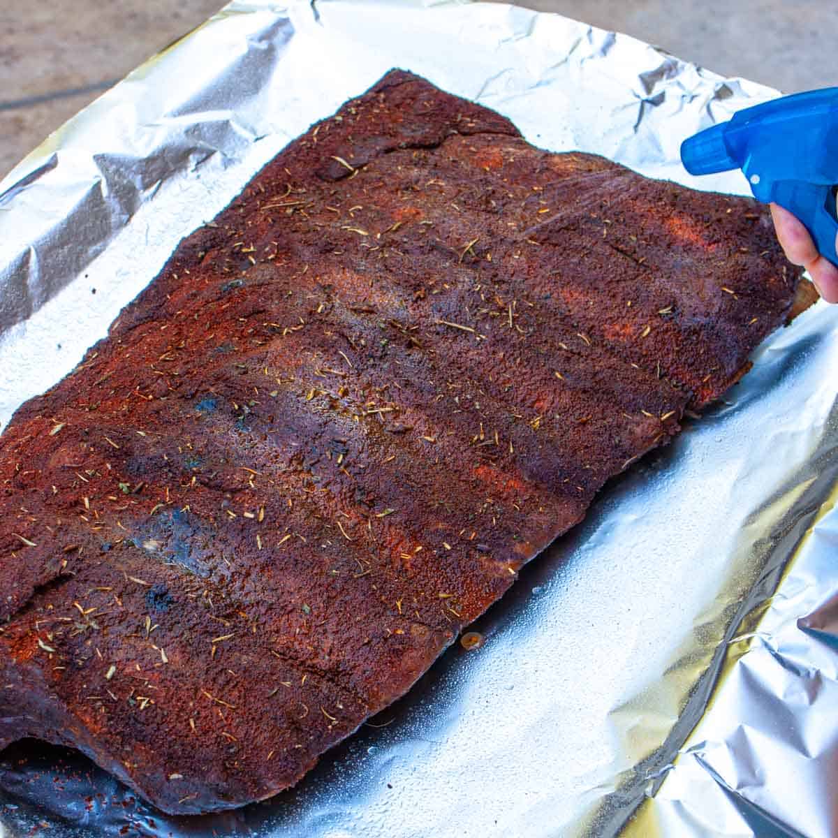 Ribs after time on smoke being spritzed with vinegar water mix just before wrapping in foil with caption.