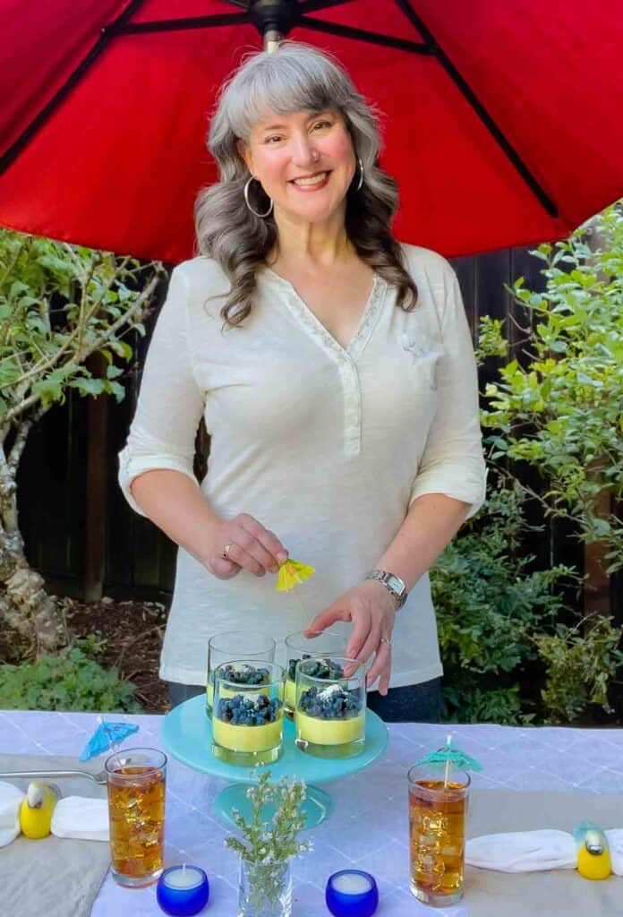 The author standing behind a table outdoors putting an umbrella in glasses of panna cotta with blueberries.