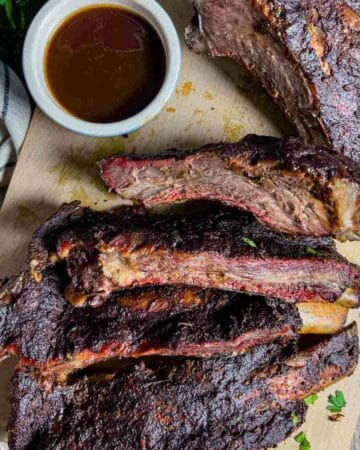 Sliced beef ribs on a cutting board with a side of barbeque sauce in a small bowl.