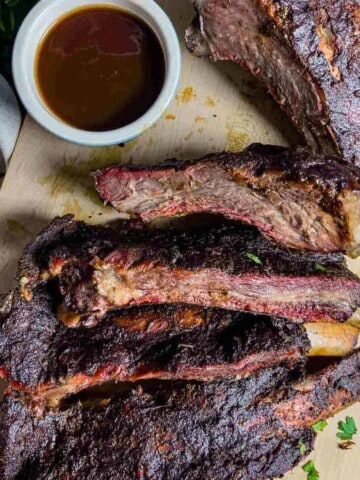 Sliced beef ribs on a cutting board with a side of barbeque sauce in a small bowl.