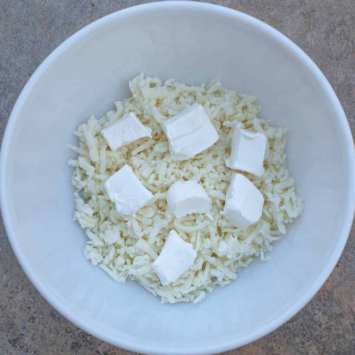 White bowl with shredded mozzarella and cream cheese chunks.