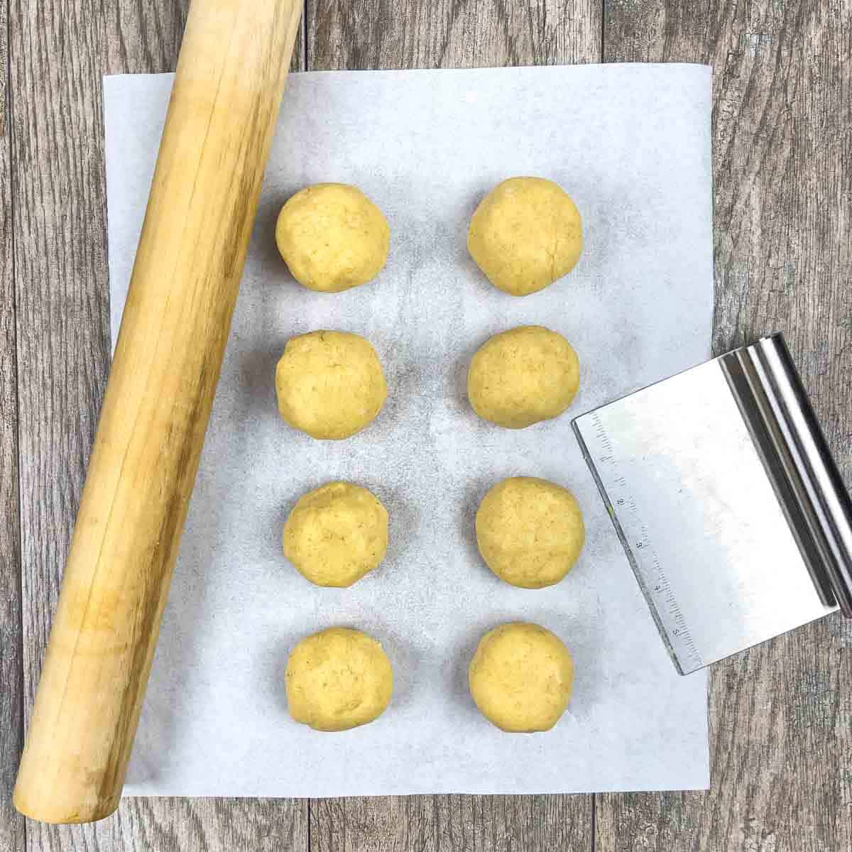 Eight small balls of dough on parchment paper with a rolling pin and a dough cutter.