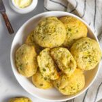 White bowl of cheddar biscuits with one in halves, with a towel and small ramekin of butter on the side.