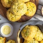 Five biscuits stacked on a board and more in a basket with one on the side broken in half next to butter in a ramekin.