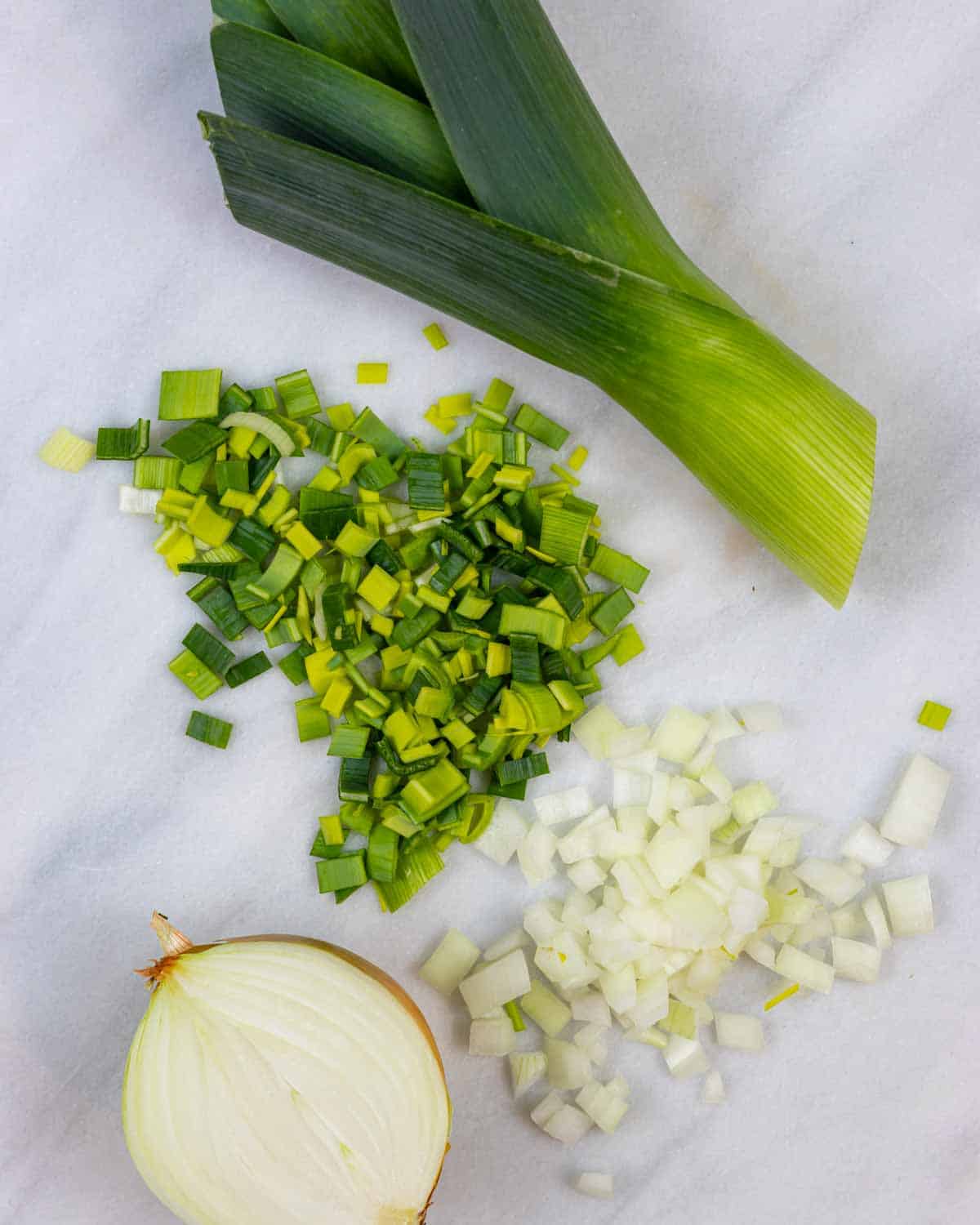 Half a leek and yellow onion on a light marble board with piles of each chopped.