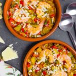 Two terra cotta cazuelas filled with Spanish vegetable stew topped with whole shrimp and manchego cheese.