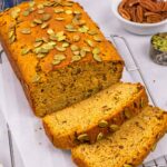 Baked loaf of pumpkin bread topped with pumpkin seeds with two slices laying in front.