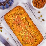 Golden baked whole loaf of keto pumpkin bread on a cooling rack with a fancy handled knife and topped with pumpkin seeds.