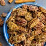 Candied pecans overflowing a blue bowl with cinnamon sticks and more pecans scattered around.