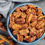 Bowl of maple bourbon candied pecans on a grey board with a grey towel.