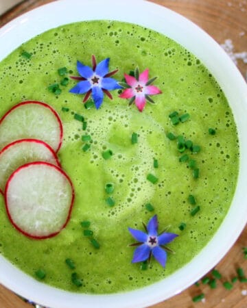 Cucumber gazpacho in a white bowl topped with radish slices, purple and pink borage flowers and fresh chopped chives.